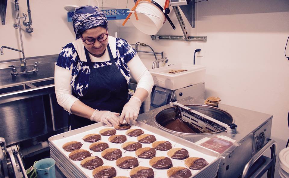 Person wearing glasses, apron, and headscarf preparing cookies in a kitchen, with trays of cookies on a table.