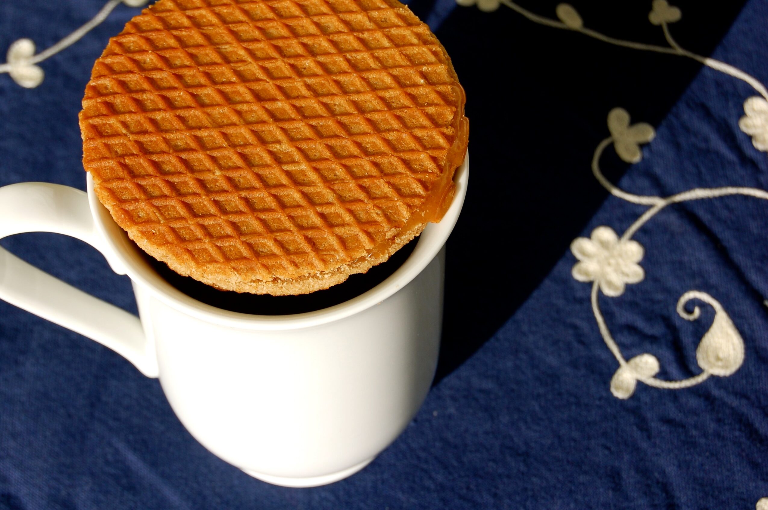 Waffle placed on top of a white mug, with a dark background and blue fabric with white floral embroidery