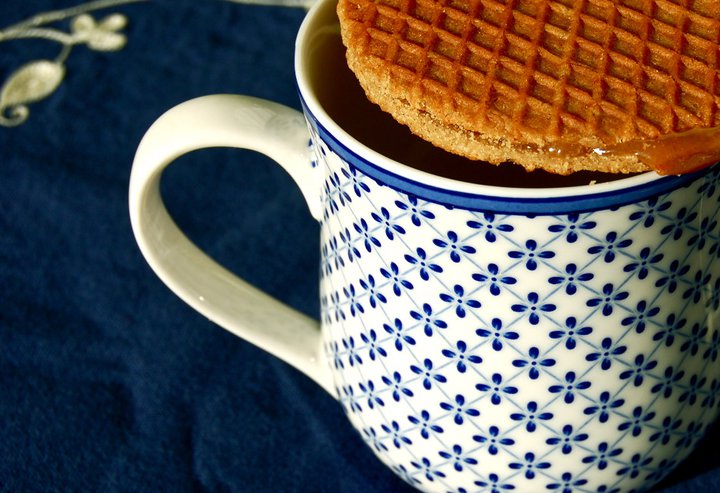 A mug with a blue floral pattern filled with a dark beverage and topped with a waffle cookie, on a dark blue surface.