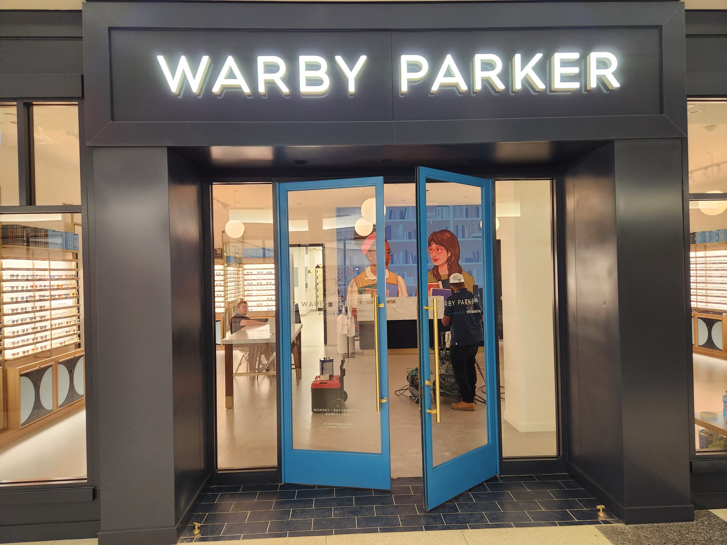 Store entrance with blue doors and two people inside, illuminated sign reads 'WARBY PARKER'.