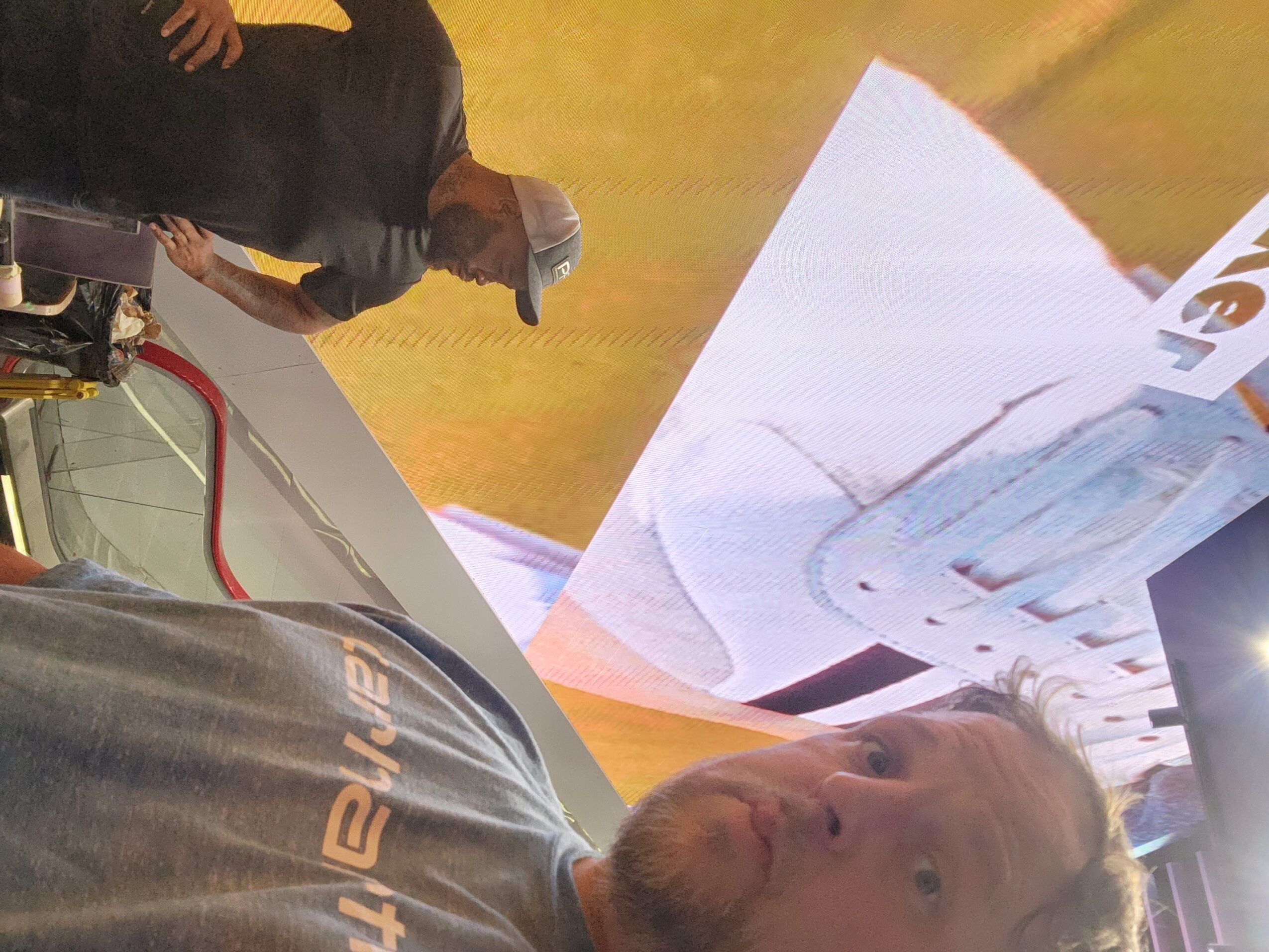 Person with light-colored hair looking at a large screen or display, with another person in the background, indoors.