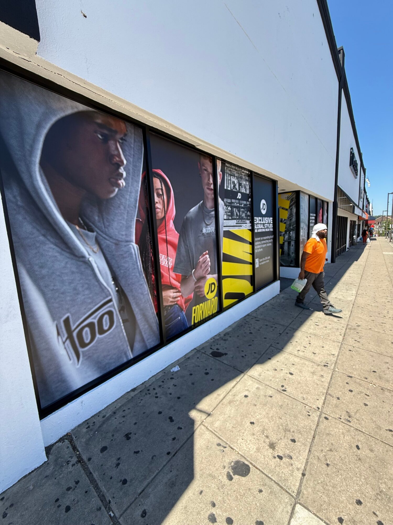 Person walking past a storefront window with advertisements, sidewalk, and shadow, on a sunny day.