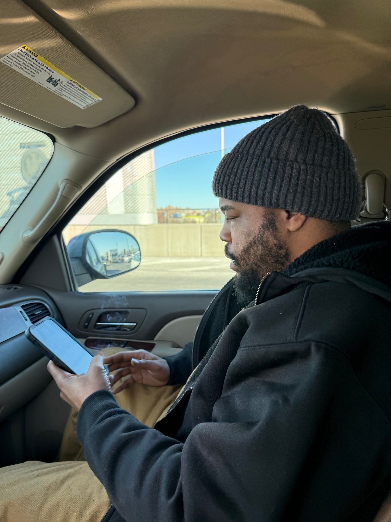 Man wearing a beanie and dark jacket sitting in car, looking outside through window, holding a phone or device.