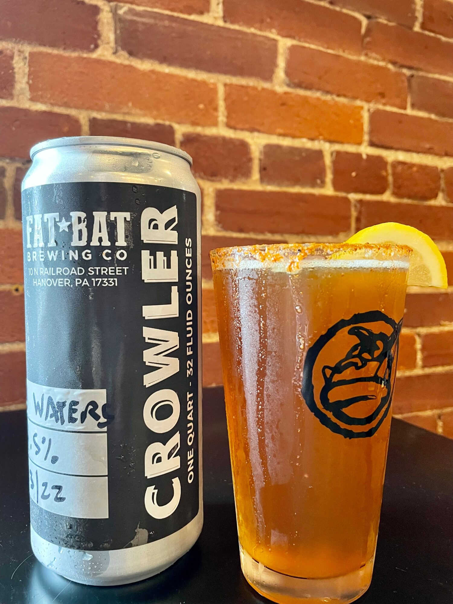 A can of Grown Lager and a glass with a lemon wedge on a dark surface against a brick wall.