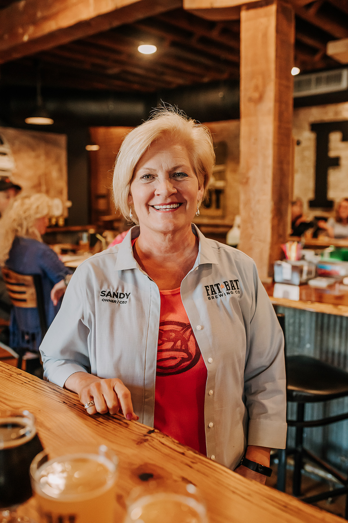 Smiling woman with short blonde hair in a white jacket standing at a bar in a restaurant or pub.