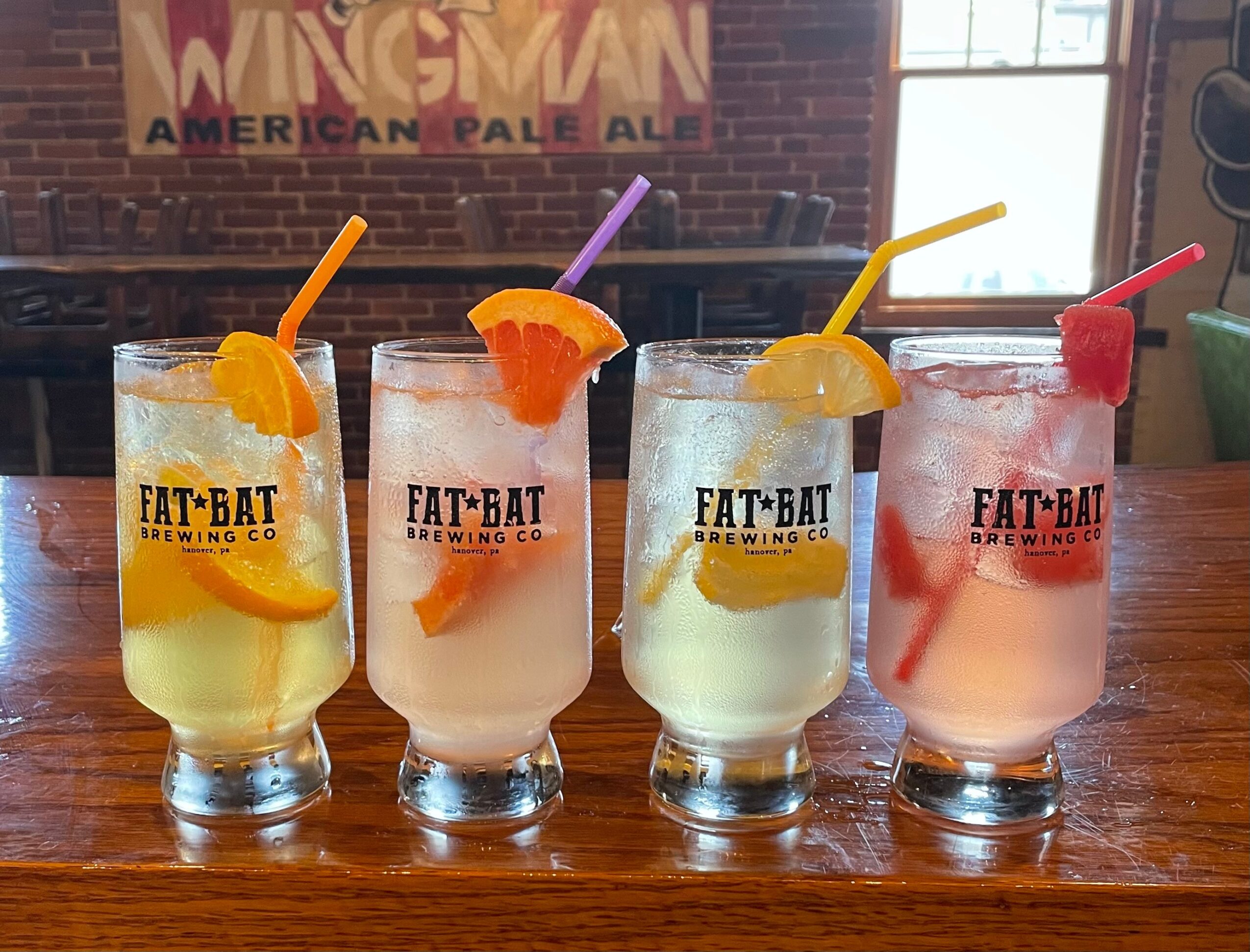 Four colorful drinks with orange, purple, yellow, and red garnishes on a wooden table, in front of a brick wall and window.