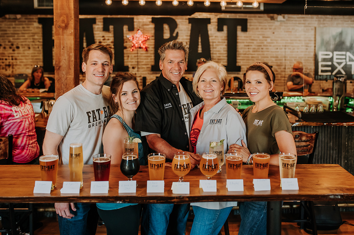 Group of five people standing behind a bar with various beers, smiling, in a brewery setting.