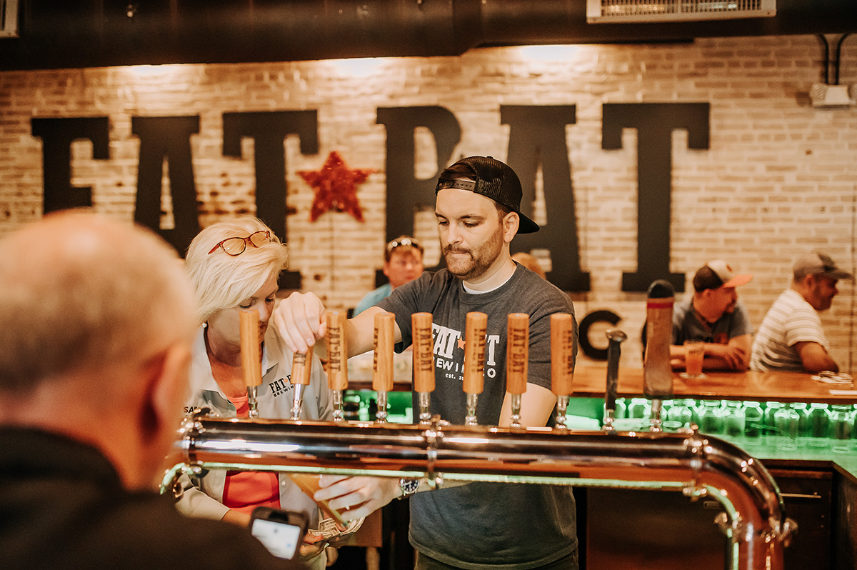 Person with a beard and headband pours drinks at a bar with a brick wall background and large text reading 'EAT BEAT'.