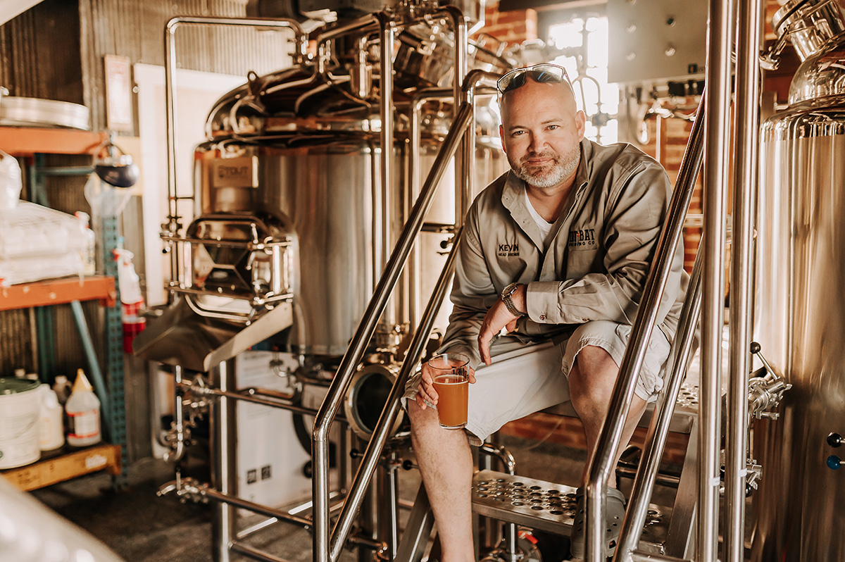 Man sitting on a chair in a brewery with brewing equipment around him, holding a glass of beer.