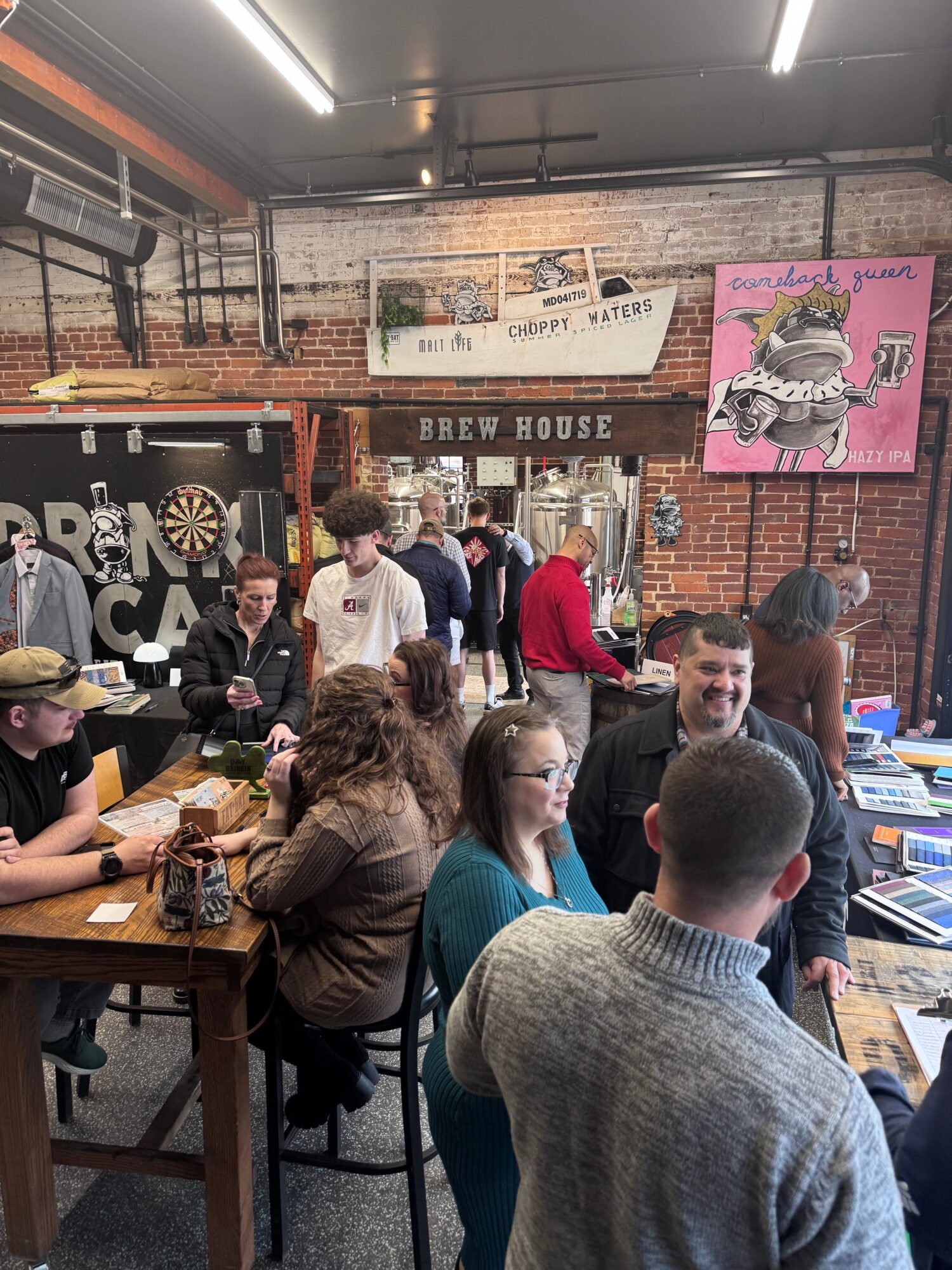 People gathered around tables in a room with brick walls and posters, engaging in conversation and activities.
