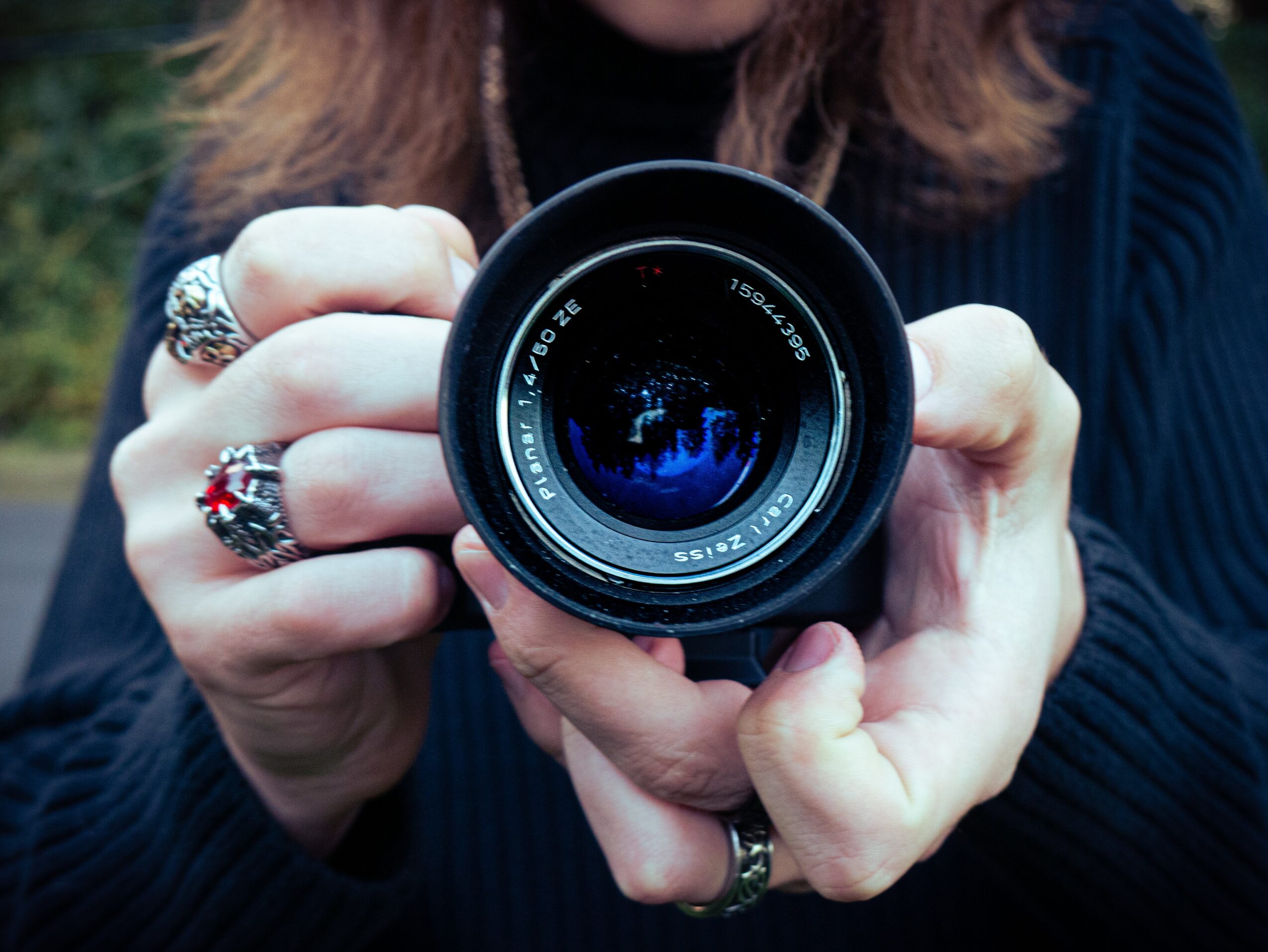 Person holding a camera lens, wearing rings, with a blurred outdoor background.