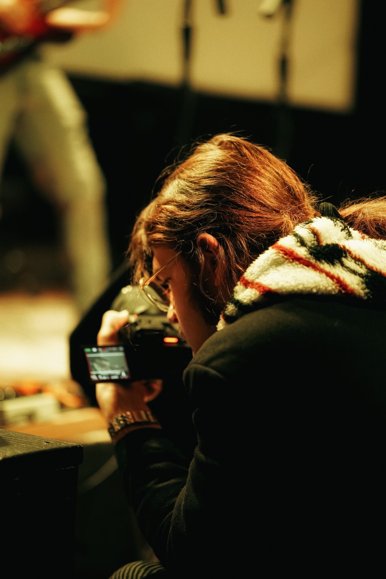 Person with brown hair and glasses looking at a camera, wearing a dark jacket and a patterned scarf, in a dimly lit environment.