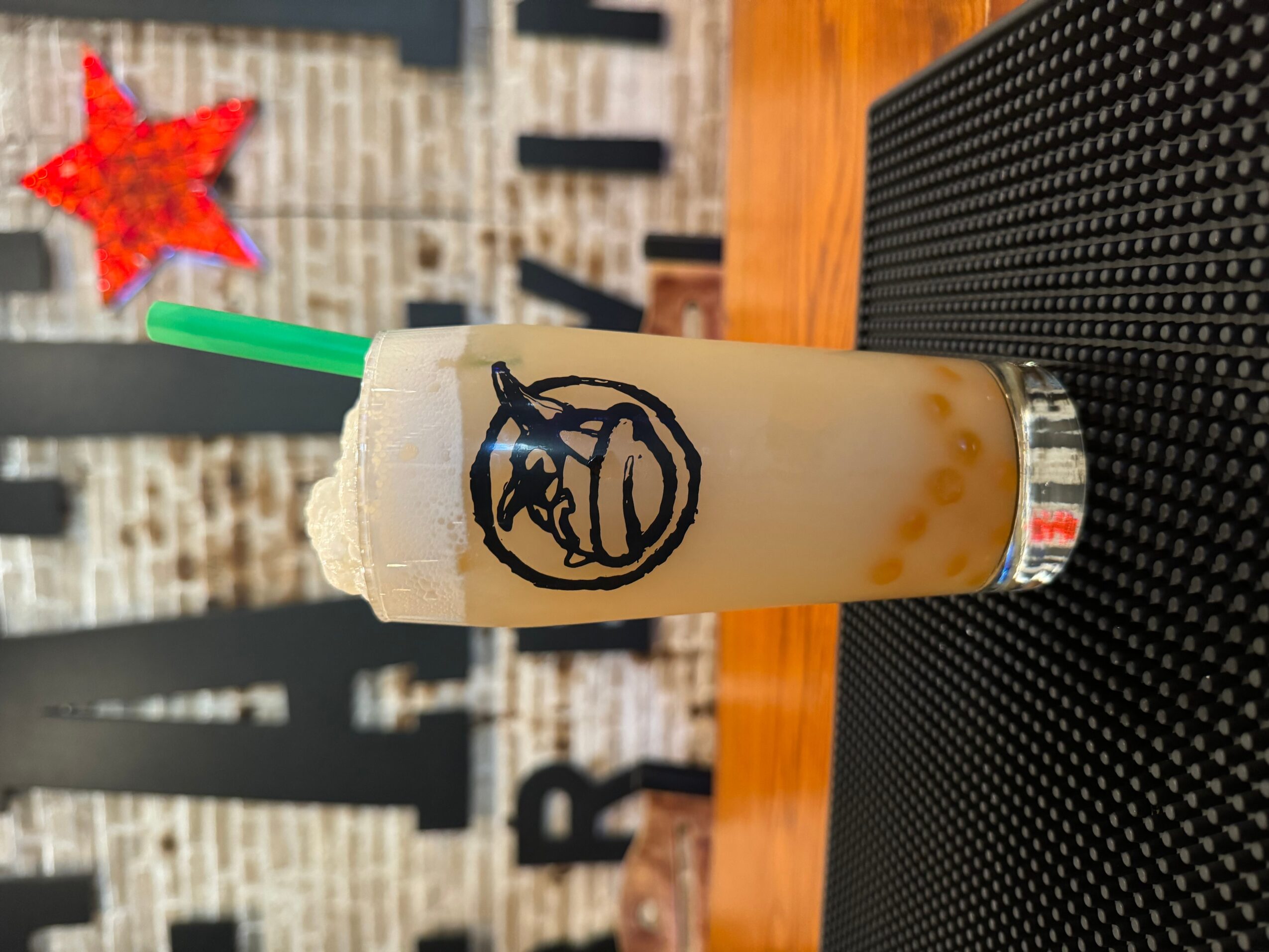 A tall glass of bubble tea with a green straw, featuring a black logo of a bear's face, on a black surface.