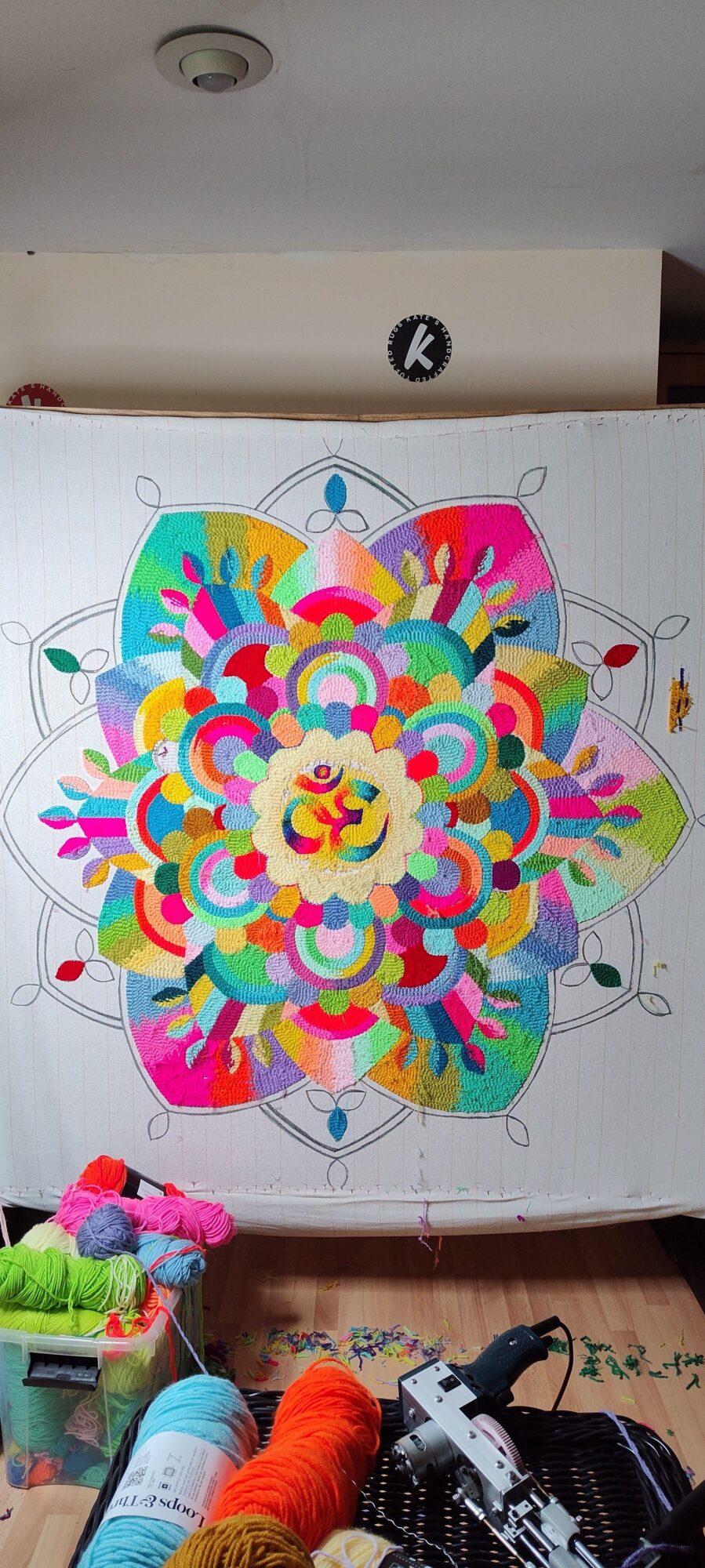 Colorful mandala with intricate patterns and symmetrical design on paper, surrounded by art supplies and a person working on it.
