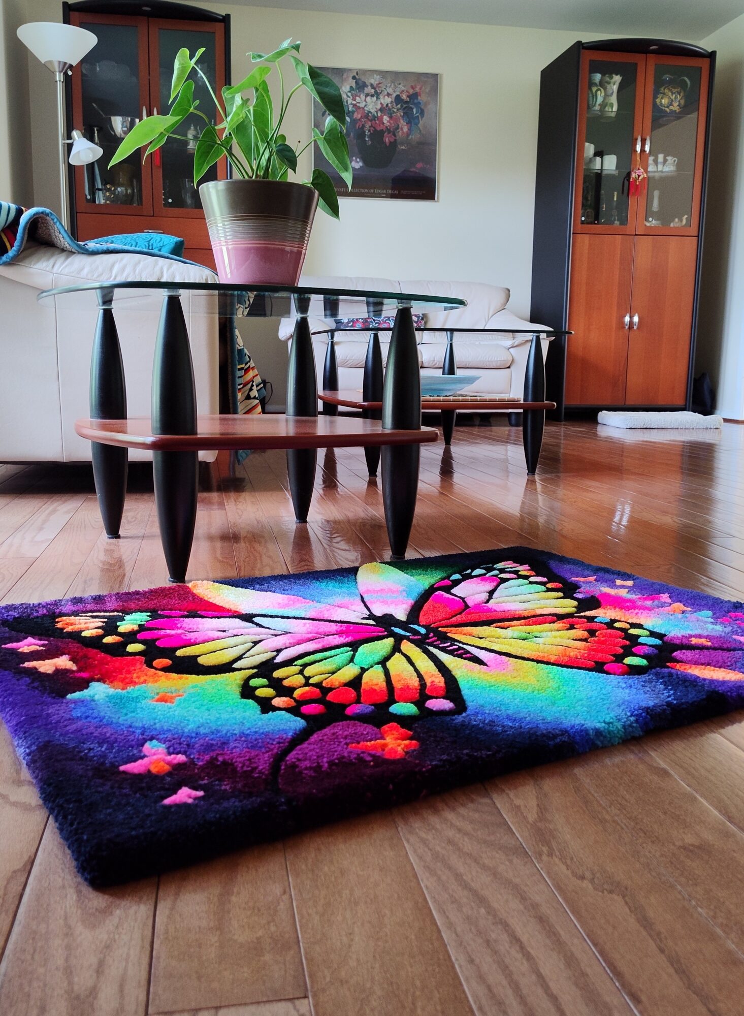 Living room with a colorful butterfly rug, glass table, chairs, and wooden cabinet, with a plant and wall art in background.