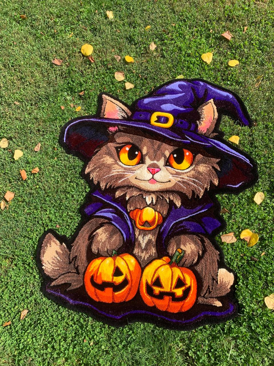 Cartoon kitten dressed as a witch with a hat, holding two carved pumpkins, on grass with fallen leaves.