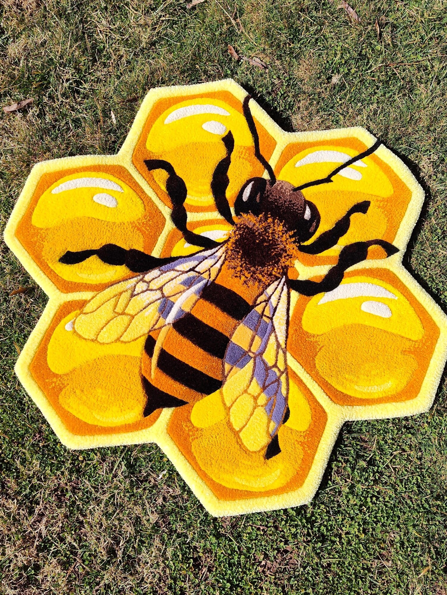 A bee illustration with yellow petals and black accents, placed on grass, with a bee in the center.