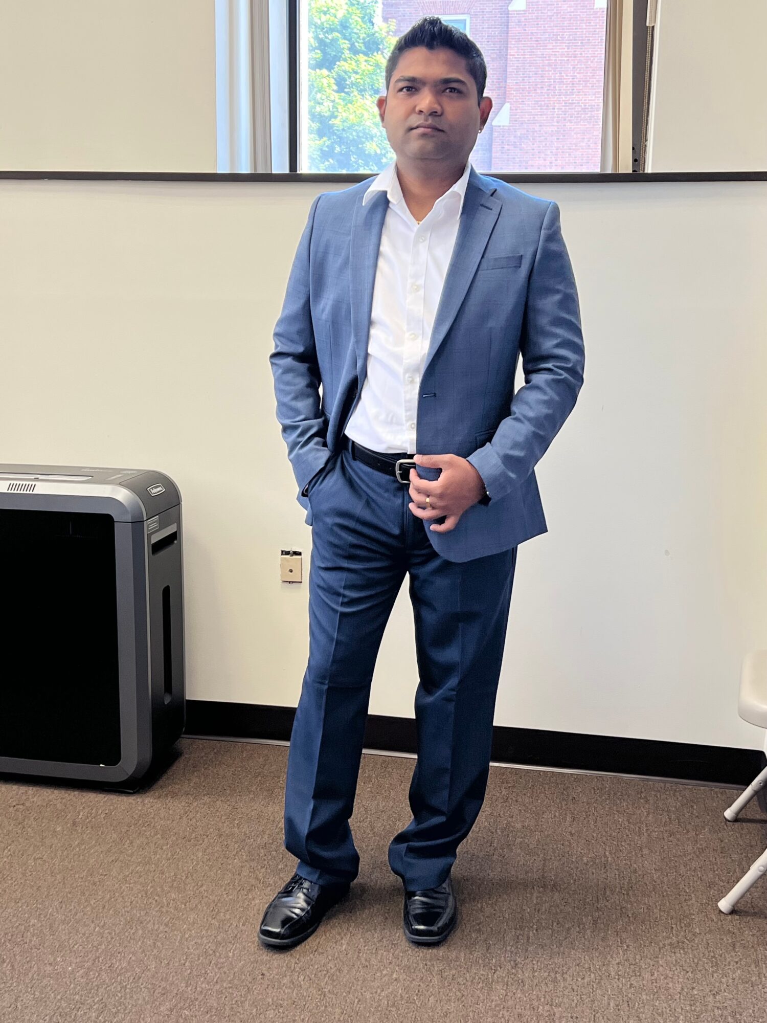 Man in a blue suit standing in an office, with a window and a computer monitor behind him.
