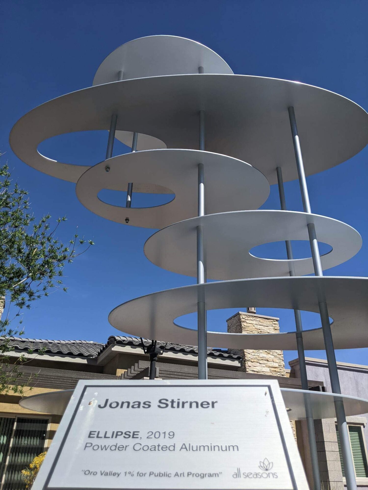 Modern sculpture with circular aluminum rings on vertical supports against a blue sky, with a building and tree below.