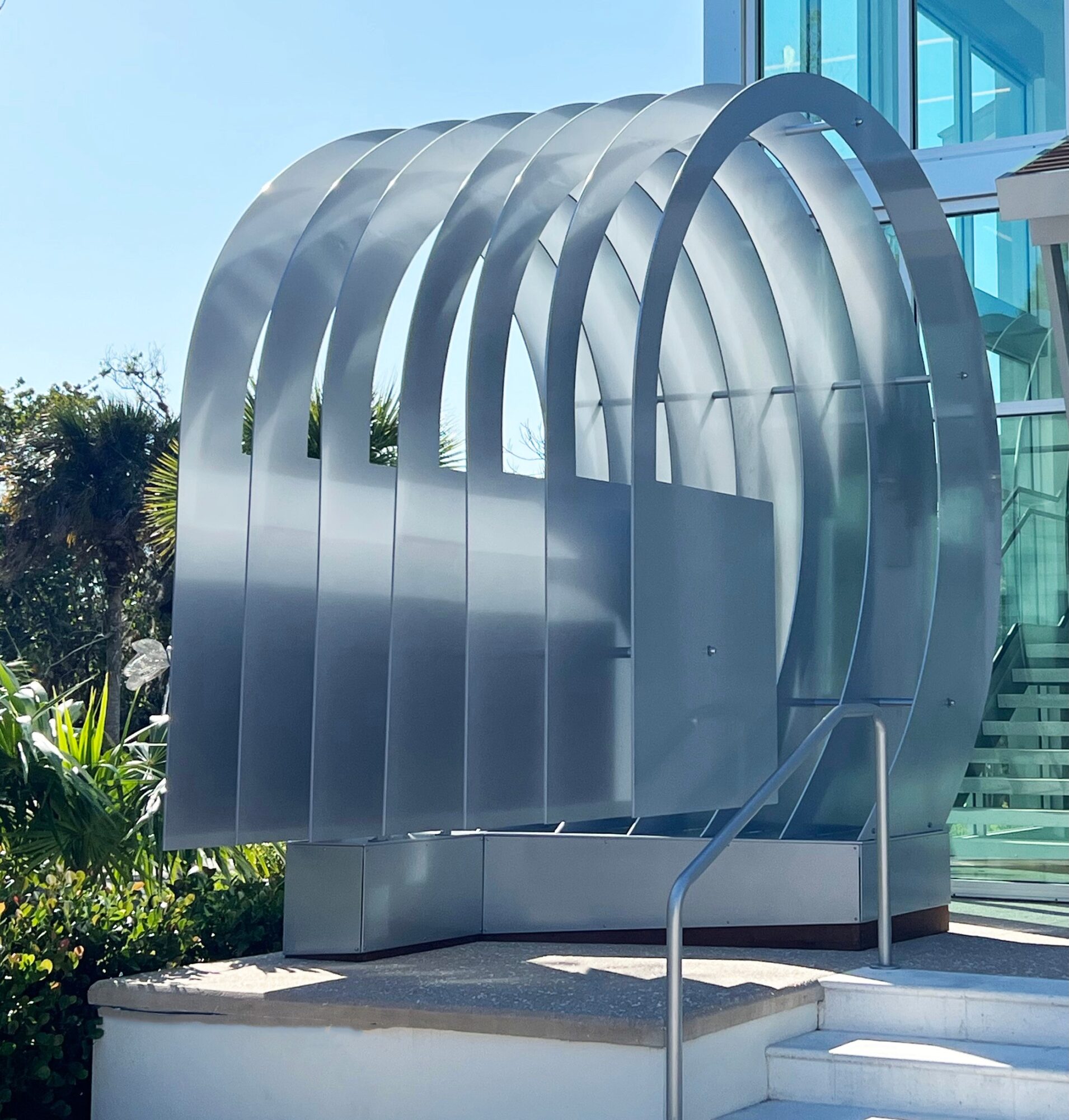 Metal sculpture with curved arches outside a modern glass building, surrounded by greenery, with stairs and a handrail nearby.