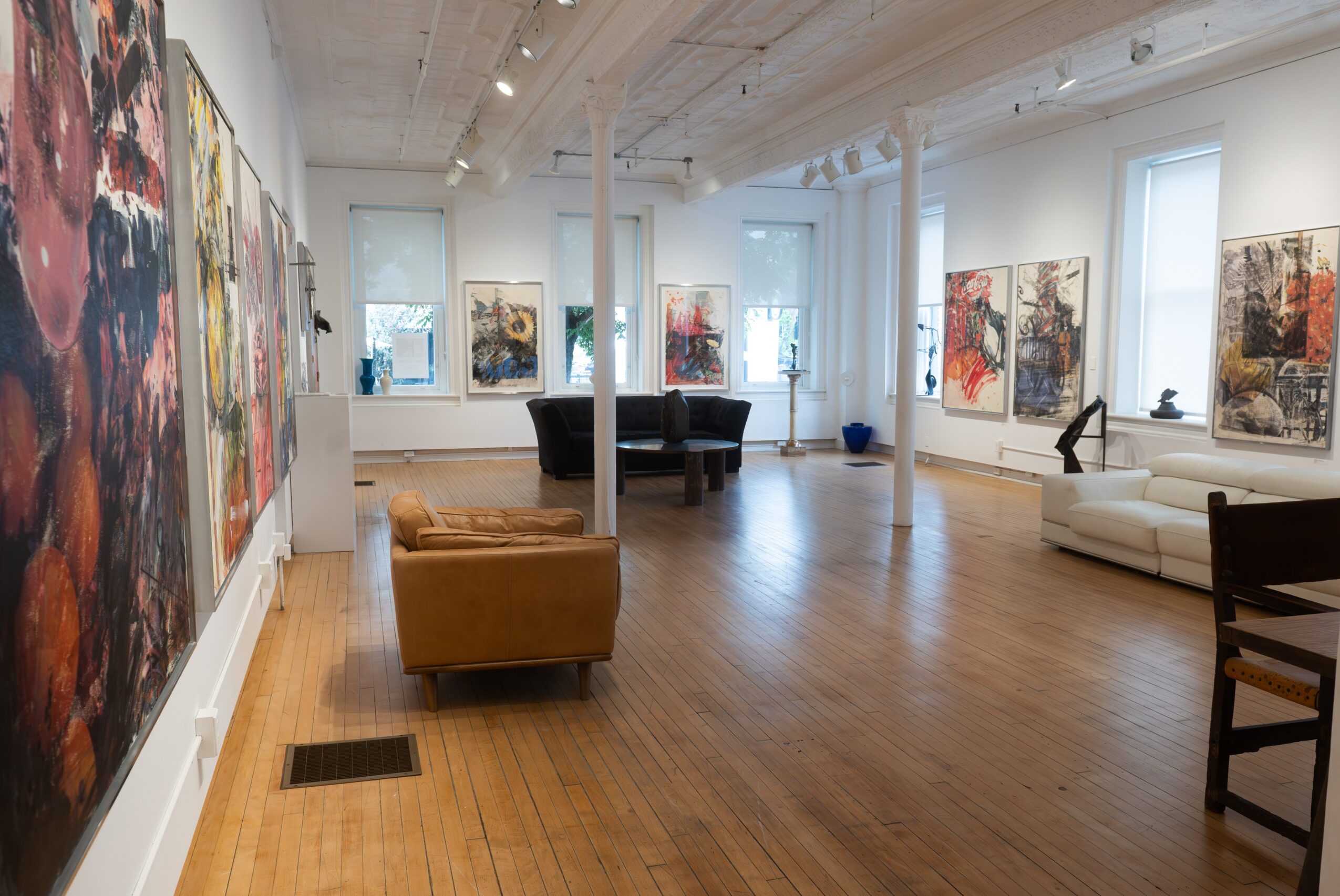 Art gallery with paintings on white walls, wooden floor, and seating areas including a brown sofa and white couch.