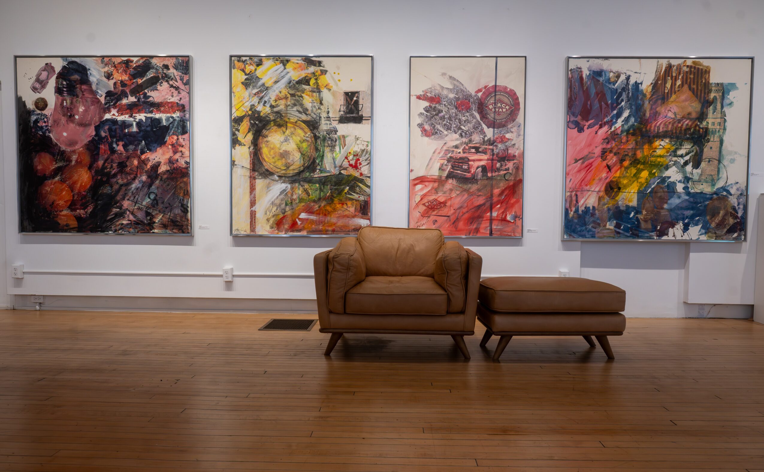 Gallery with four abstract paintings on the wall, a brown sofa, and an ottoman in front, wooden floor, white walls.