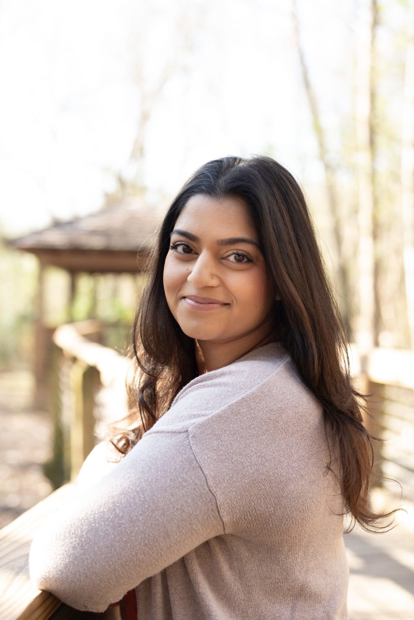 Young woman with long dark hair smiling outdoors, wearing a light-colored sweater, with trees and a wooden structure in background.