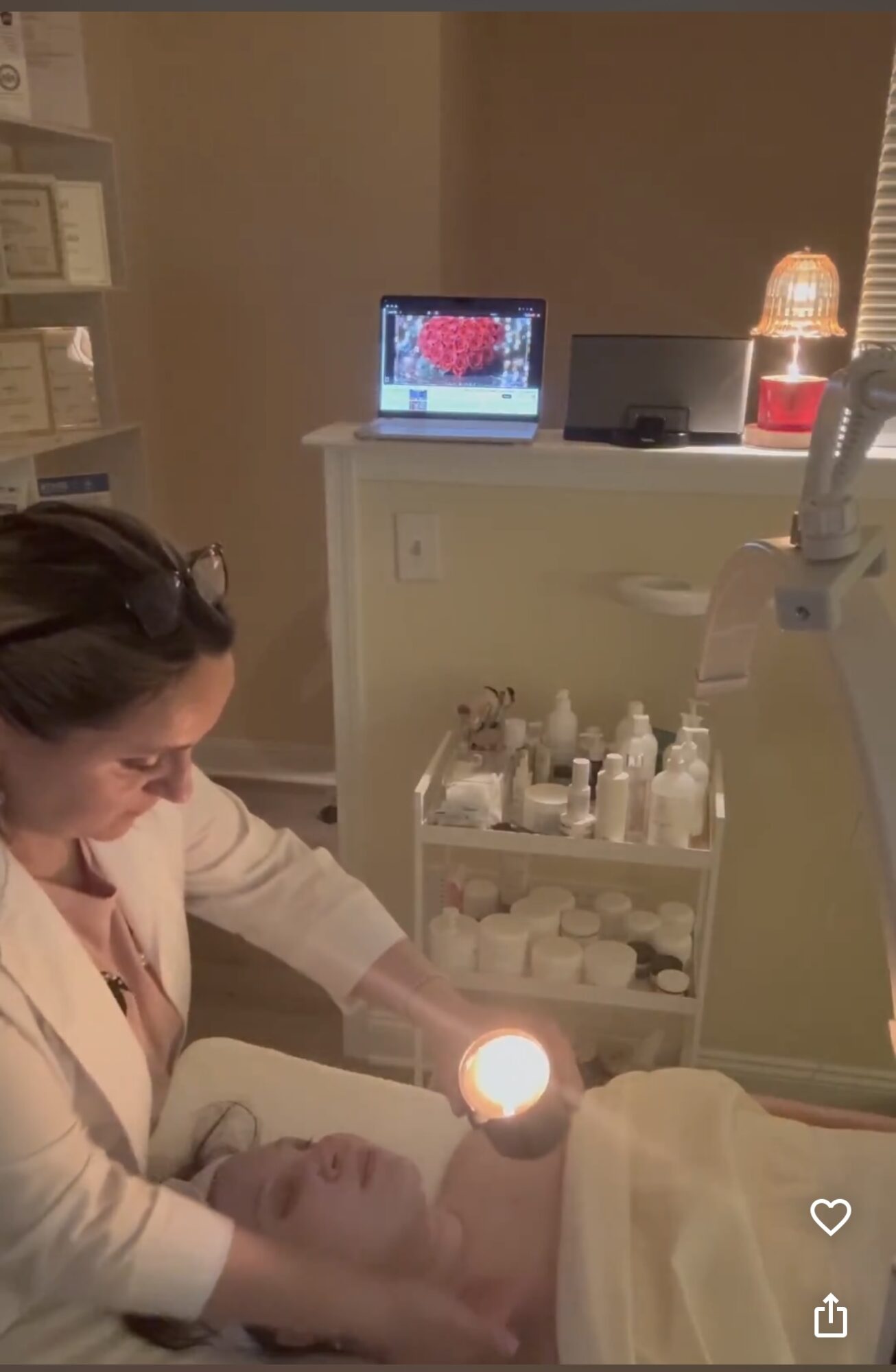A woman in a clinical setting examines a patient lying on a bed with a bright light, medical equipment nearby, and a monitor in the background.