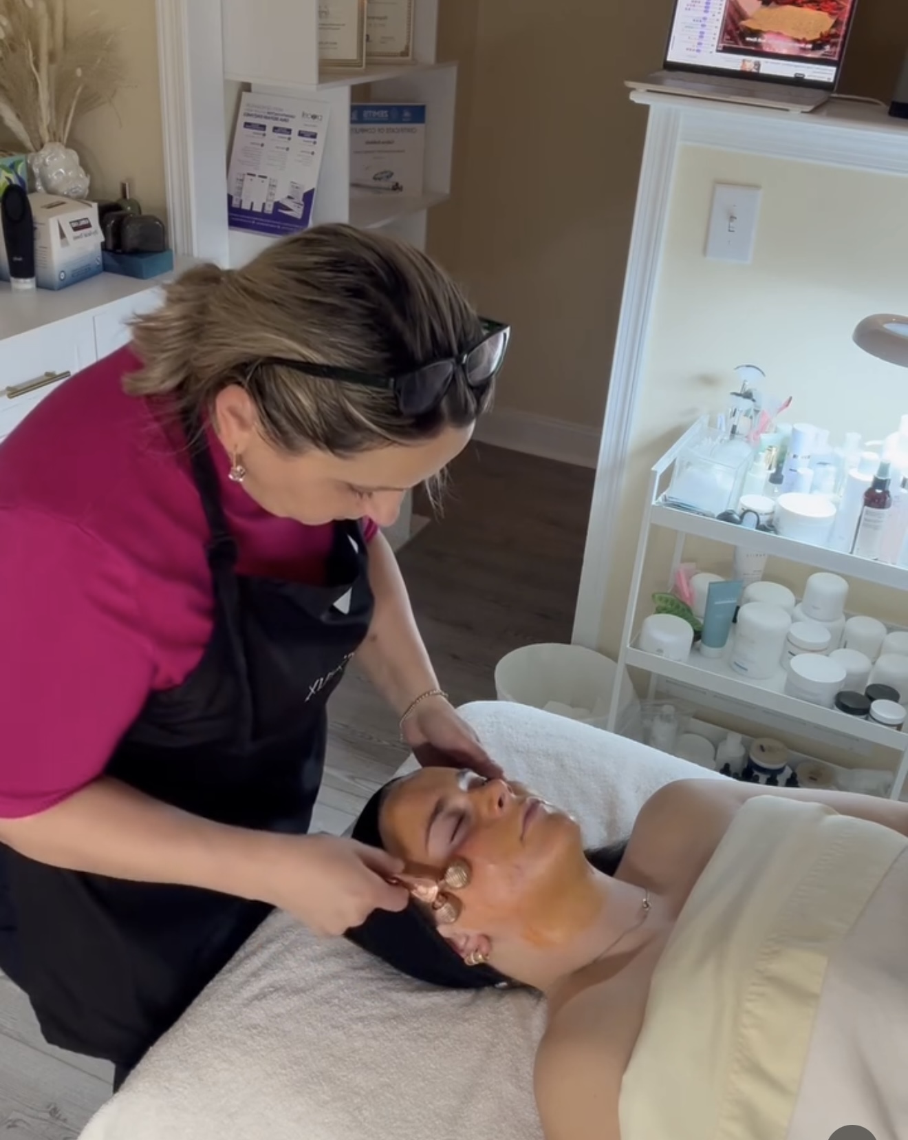 A woman receives a facial treatment from a professional in a spa or clinic setting, with skincare products nearby.