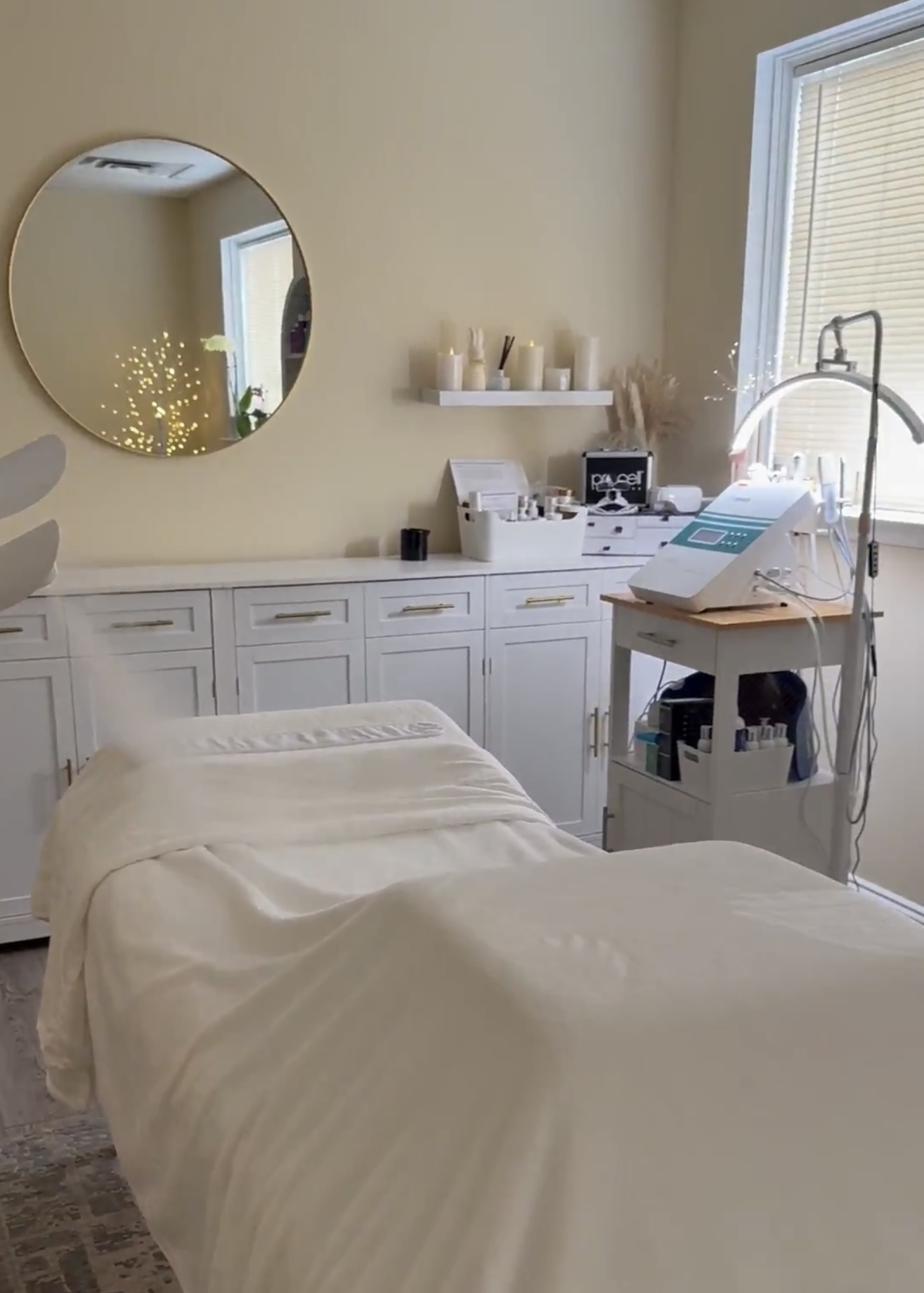 A treatment room with a bed, mirror, shelves, and equipment near a window, in a neutral-colored space.