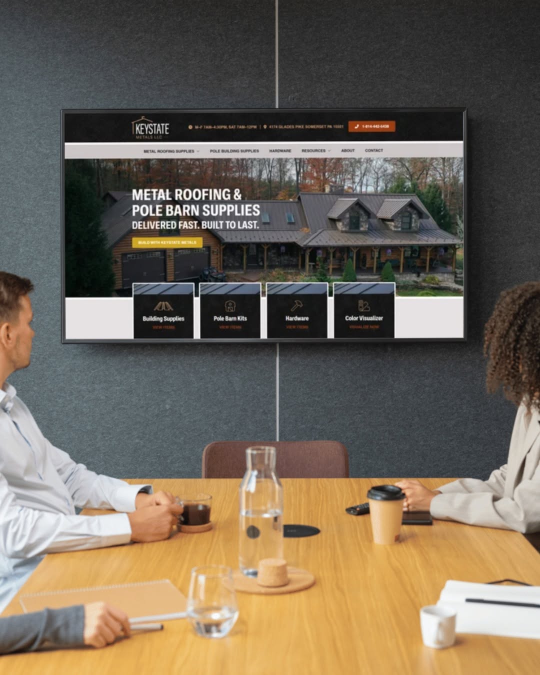 Two people sit at a conference table with drinks, facing a large screen displaying a website about metal roofing and supplies.