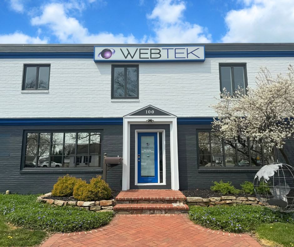 Two-story building with a sign reading 'WEBTEK' and a blue door, surrounded by a garden with a flowering tree.
