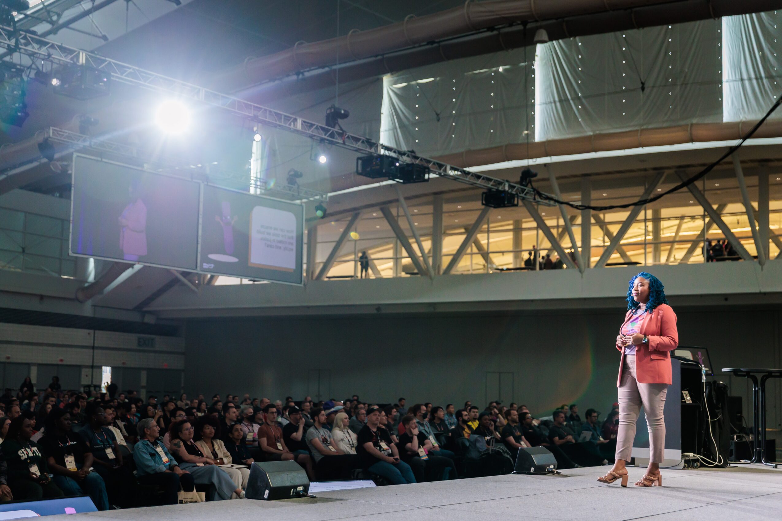A woman in a red blazer and beige pants speaks on stage to a large audience in a conference hall.