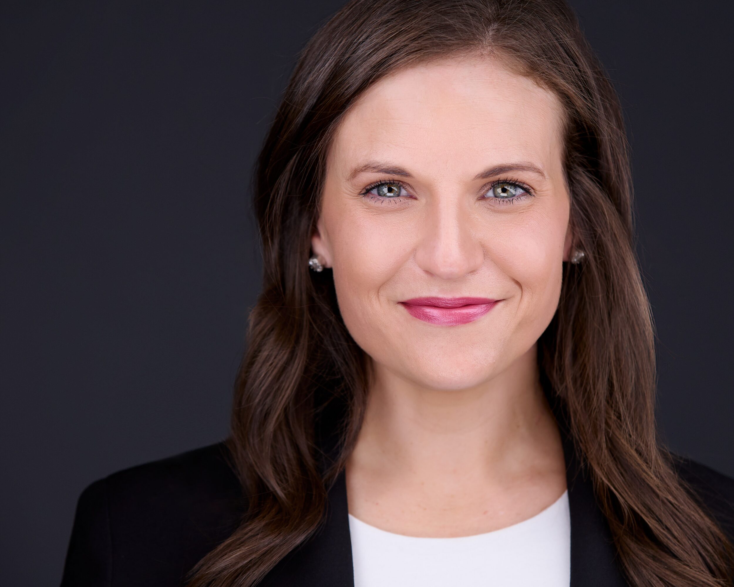 Portrait of a woman with brown hair, blue eyes, wearing a black blazer and white top, smiling against a dark background.