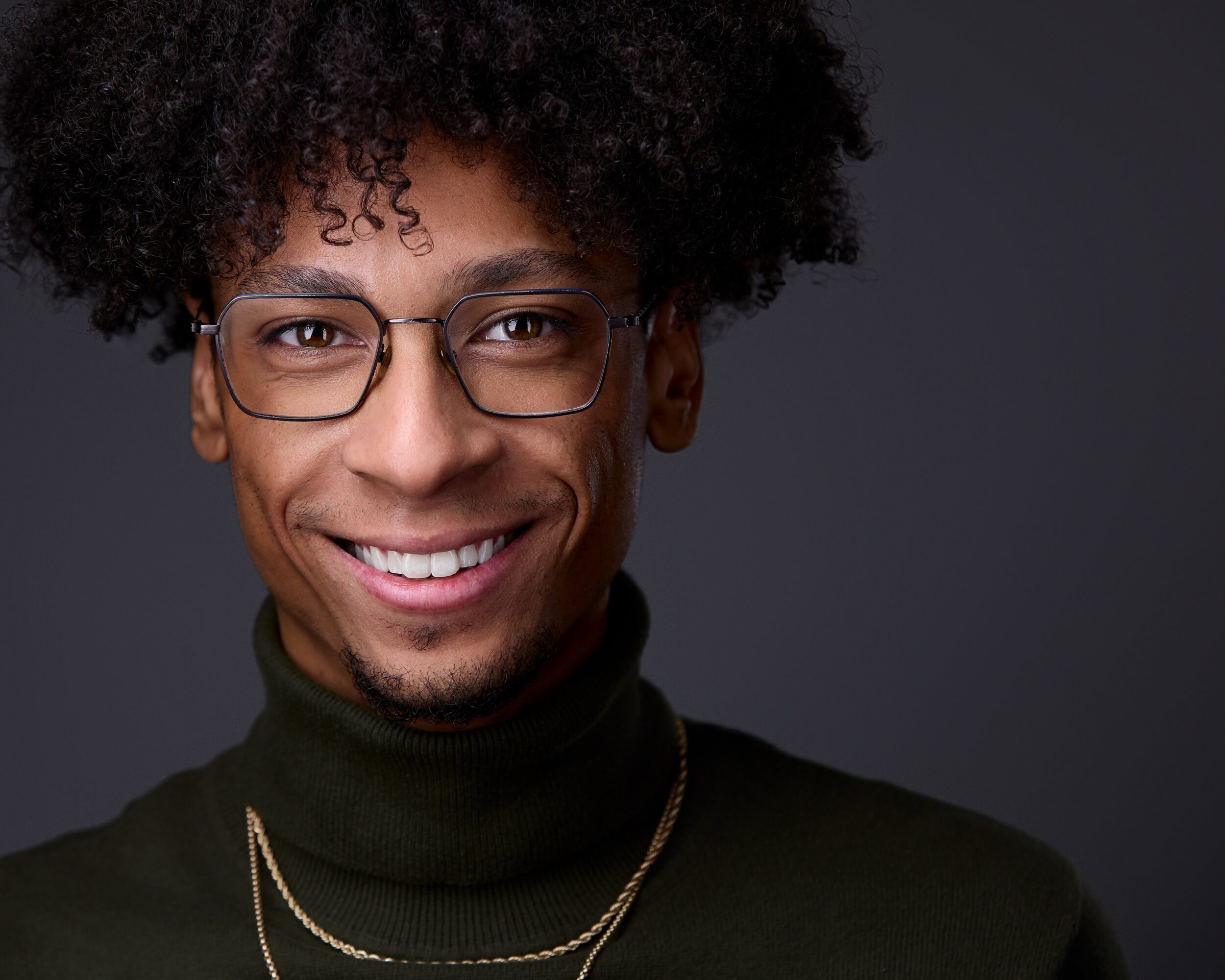Person with curly hair, glasses, smiling, wearing a black turtleneck and gold necklace, against a dark background.
