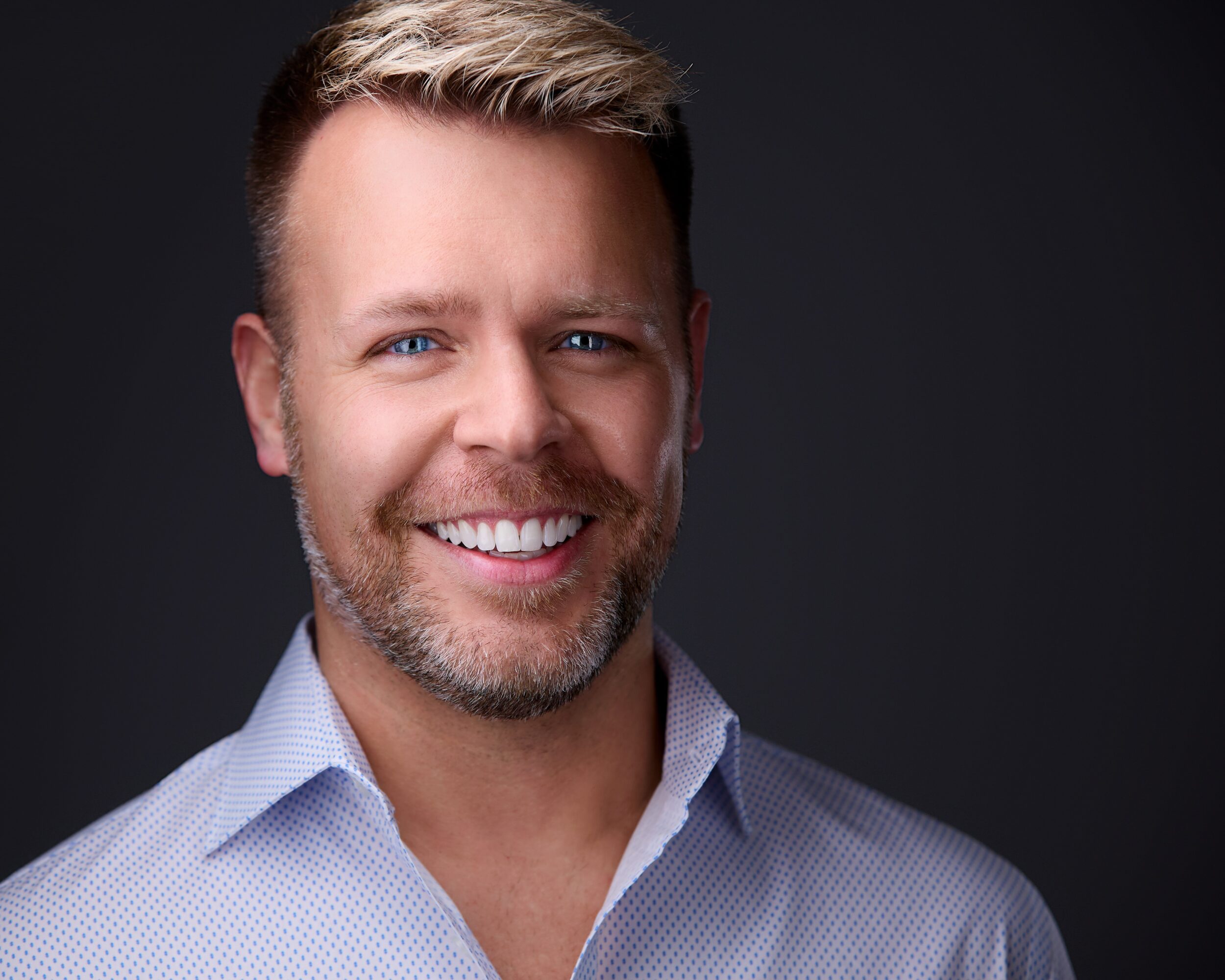 Smiling man with short hair and beard, wearing a light blue shirt, against a dark background.