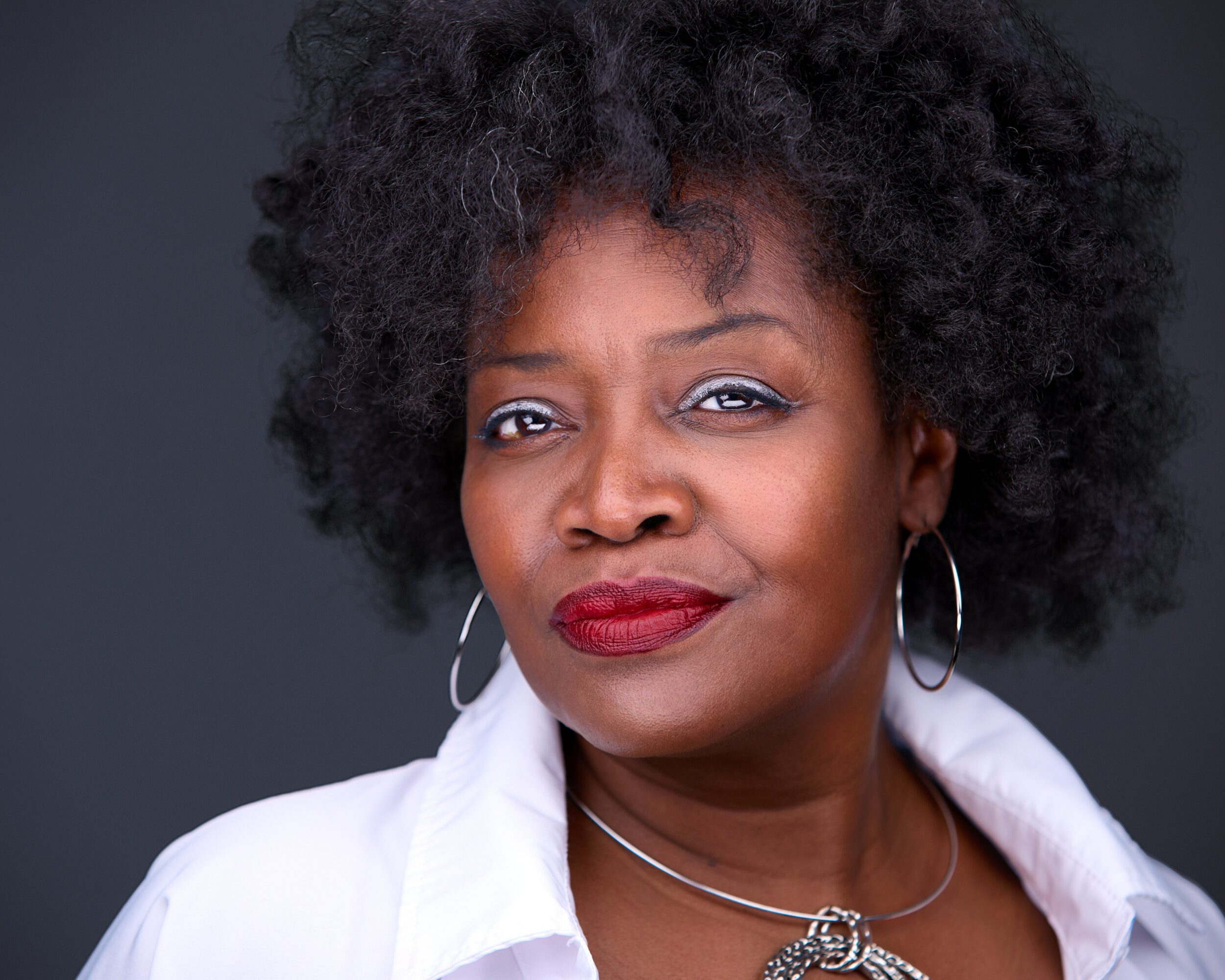 Woman with curly hair, wearing hoop earrings and a white shirt, winks at the camera.