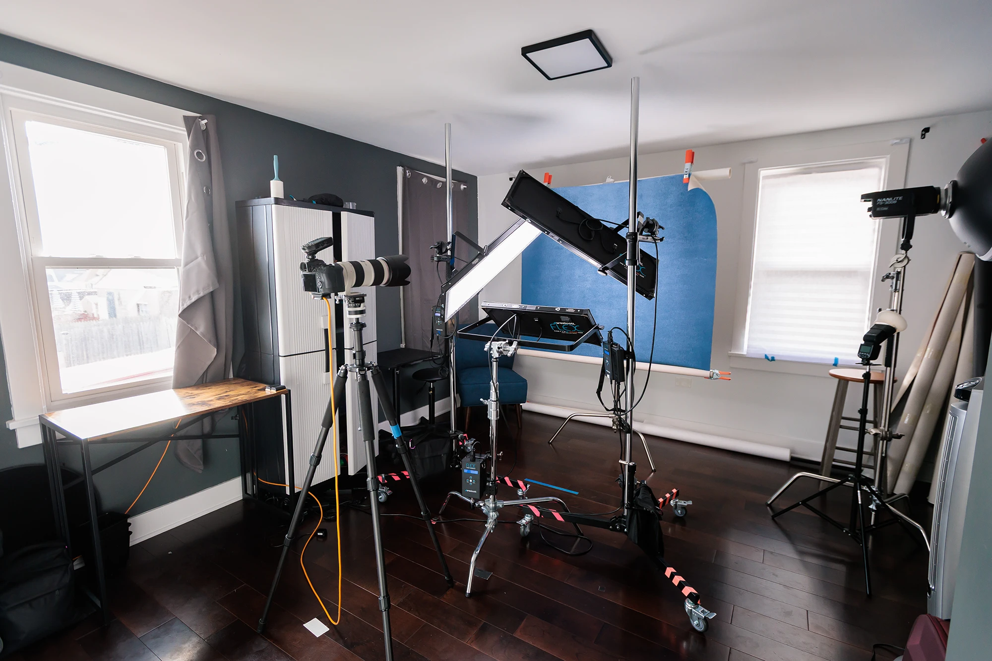 Room with telescopes, camera, and equipment setup near windows, dark hardwood floor, and blue backdrop on wall.