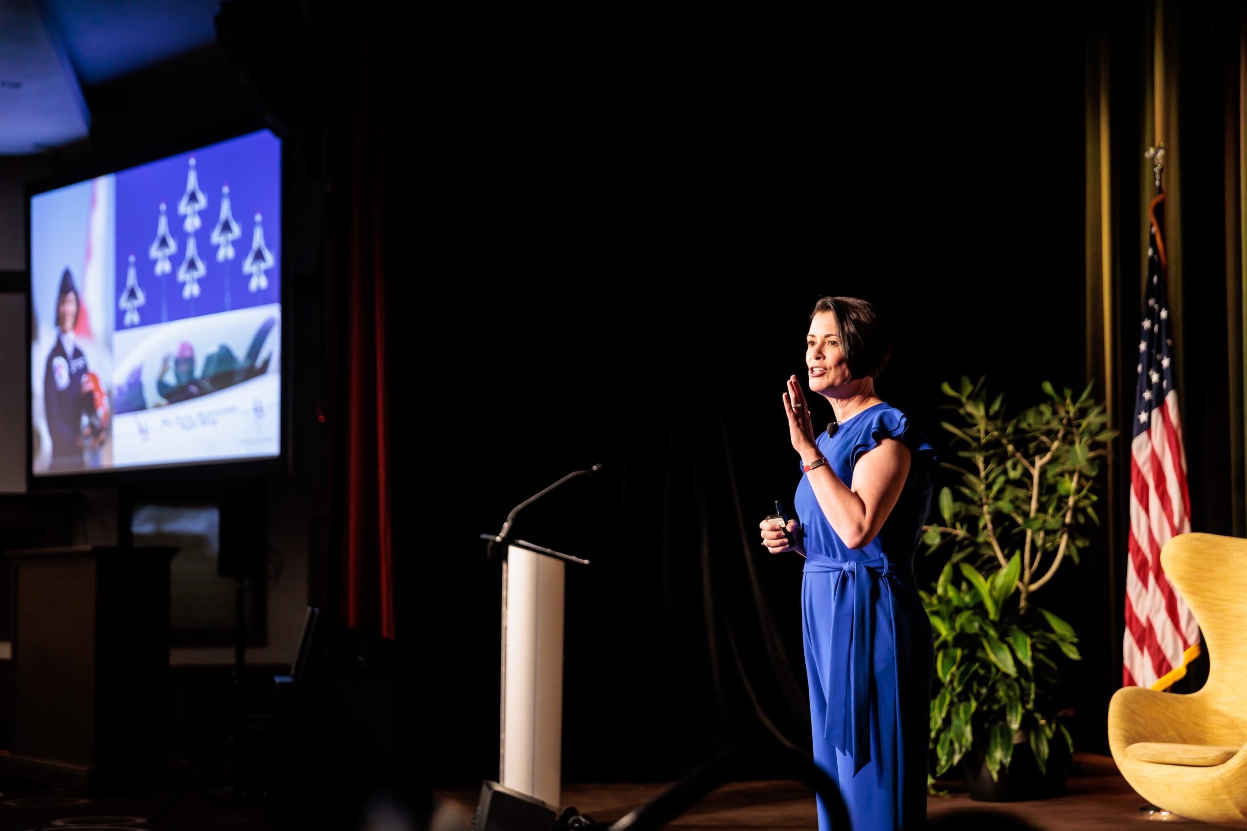 Young girl in a blue dress performing on stage with a microphone, decorated with plants and an American flag, in a dark auditorium.