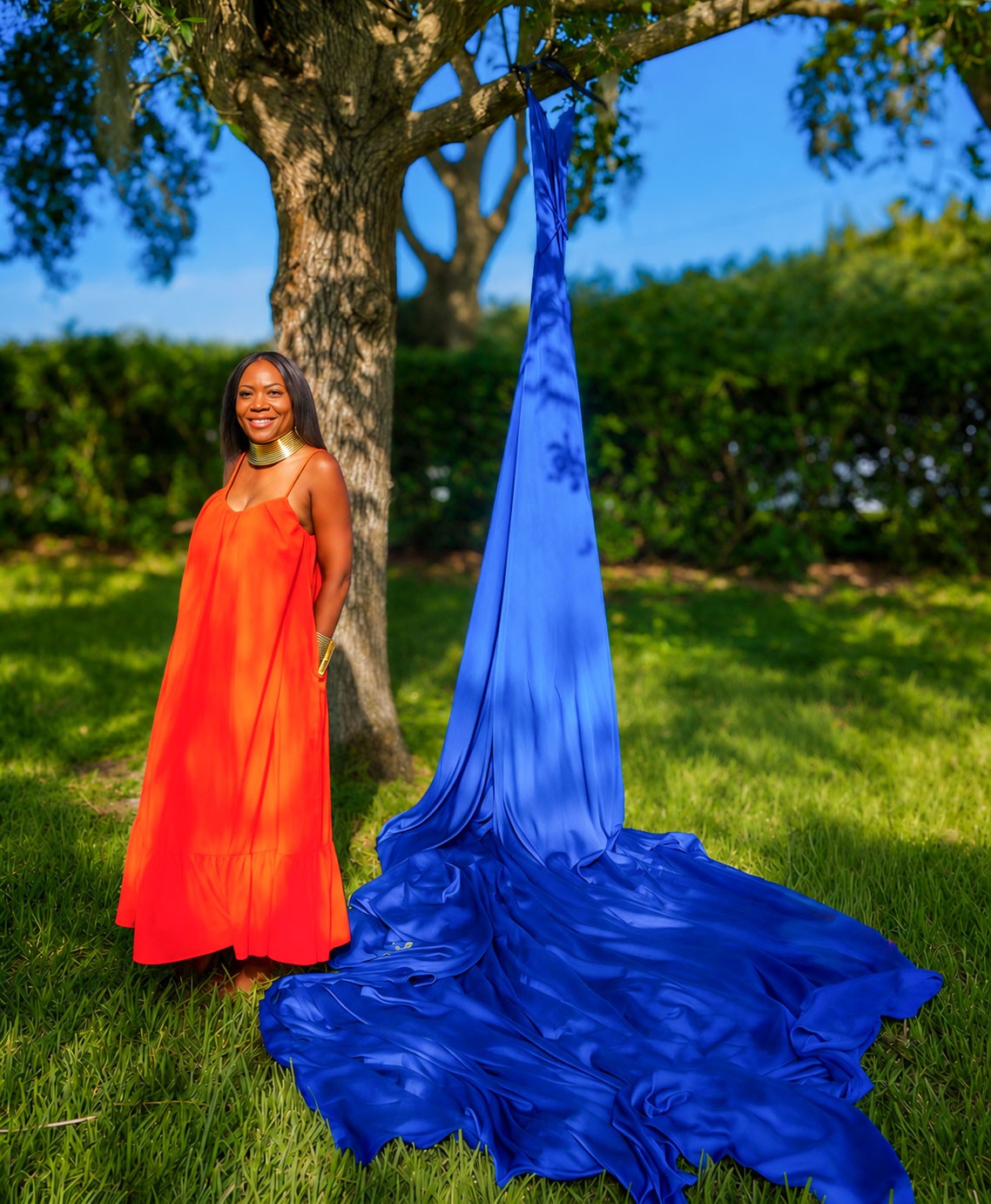 Woman in an orange dress standing outdoors next to a tree with blue fabric hanging and pooling on grass.