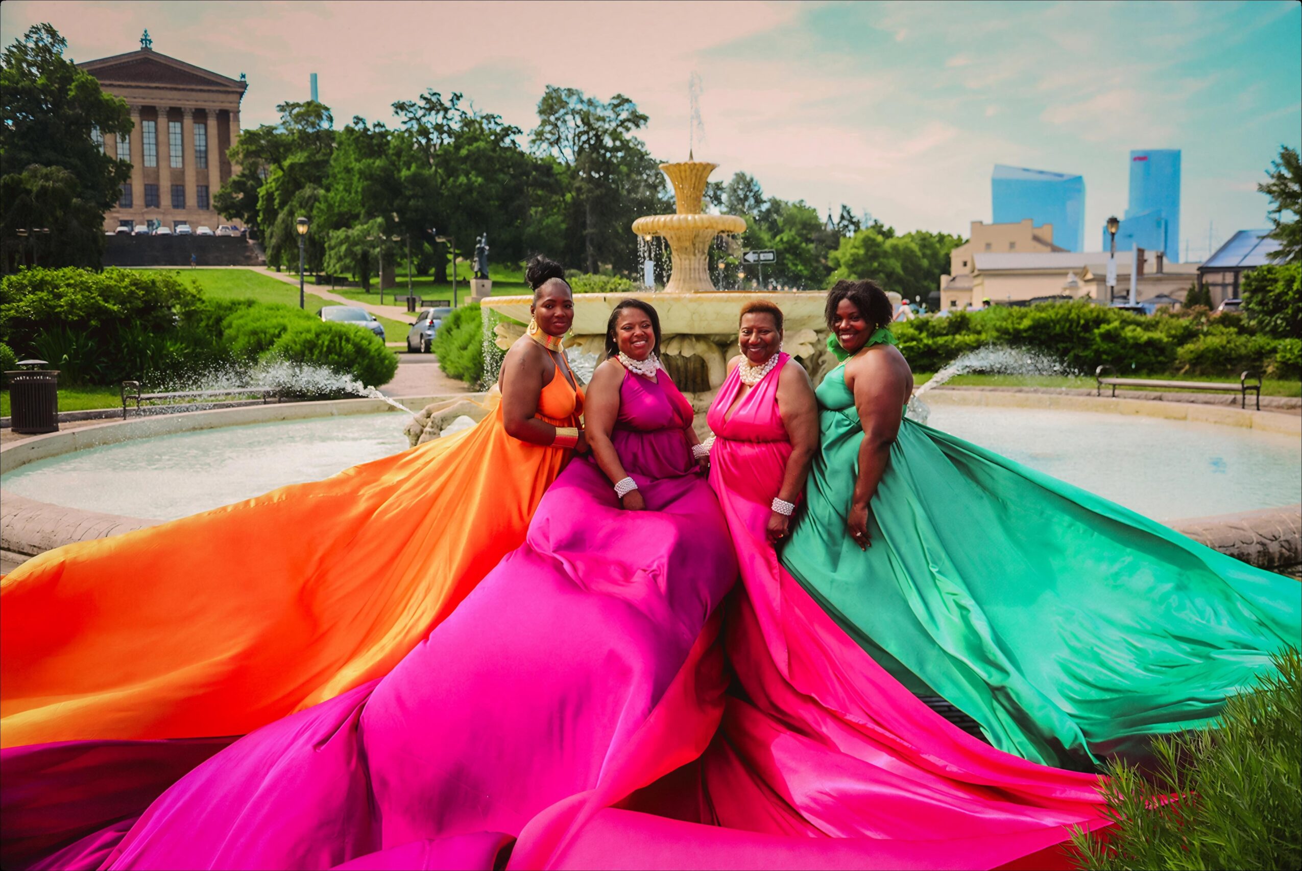 Four women in colorful dresses pose in a park with a fountain and city buildings in the background.