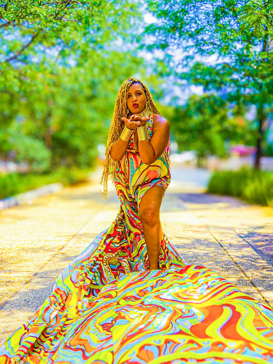 Woman with long braided hair in a colorful dress standing on a pathway surrounded by green trees, with a flowing patterned train.