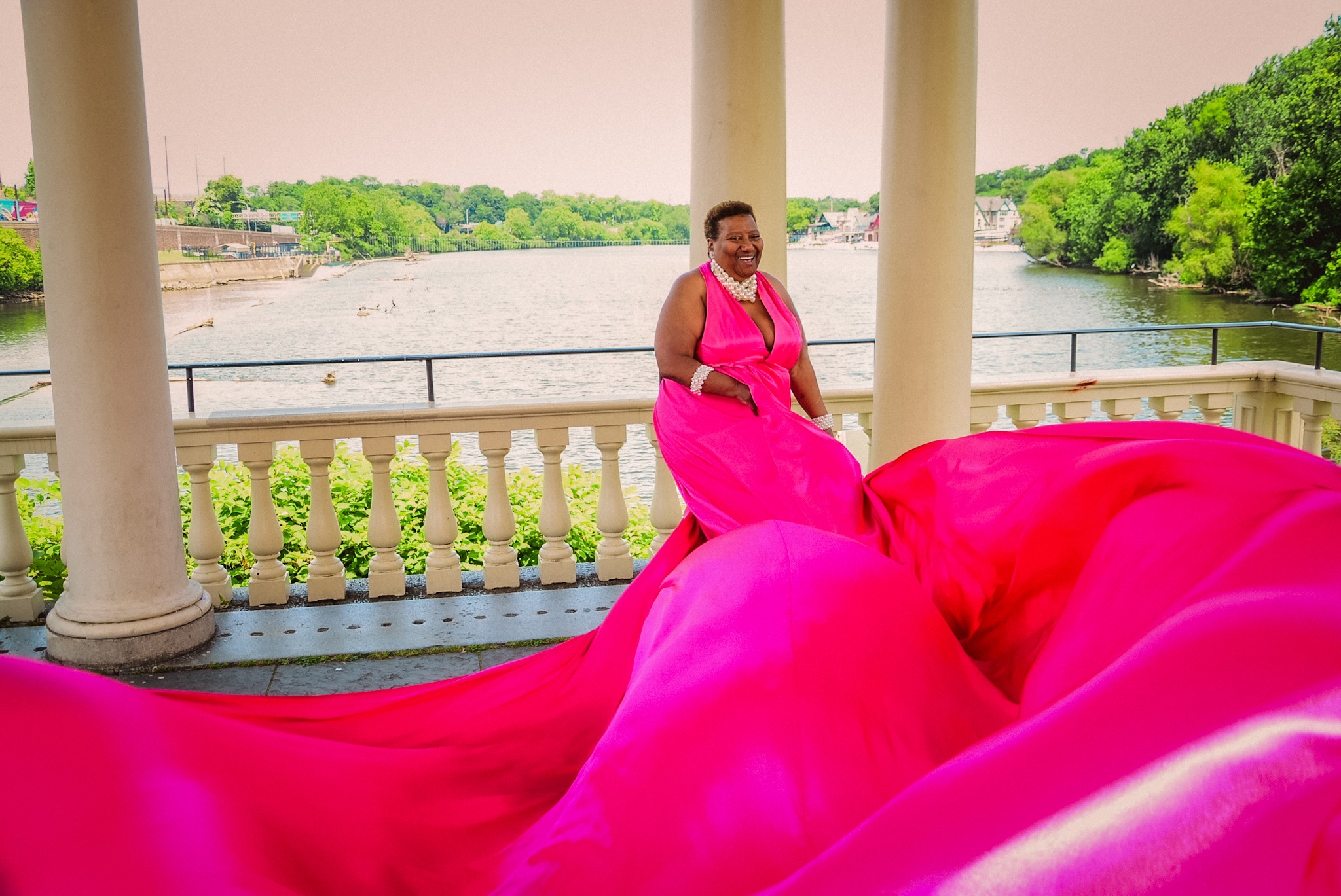 Woman in a pink dress standing on a balcony overlooking a river with trees, with a flowing pink fabric in the foreground.