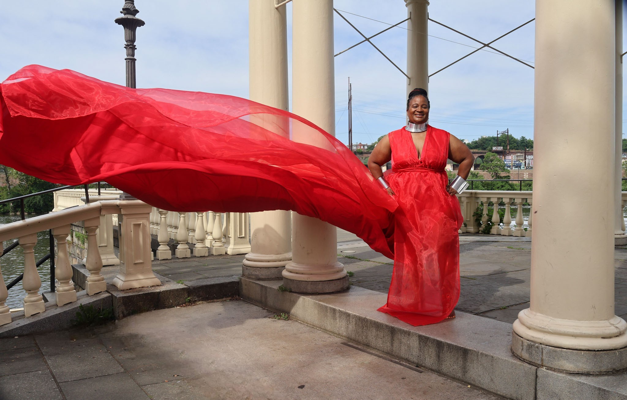 Person in a red dress standing outdoors on a stone platform with columns and a railing, holding flowing fabric.