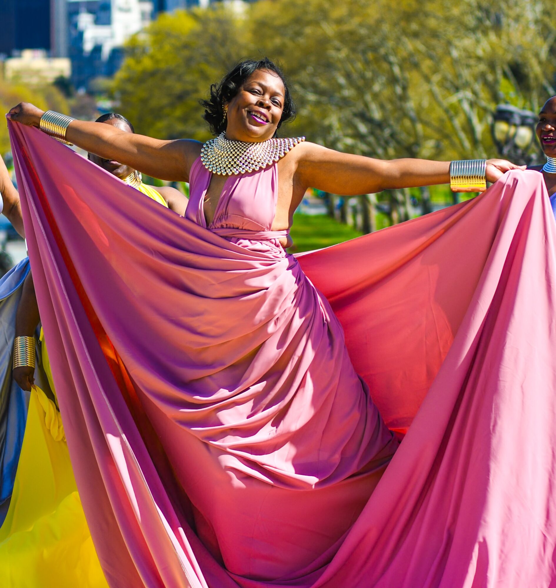 Woman in pink dress holding fabric outdoors, smiling, with trees and blue sky in background.