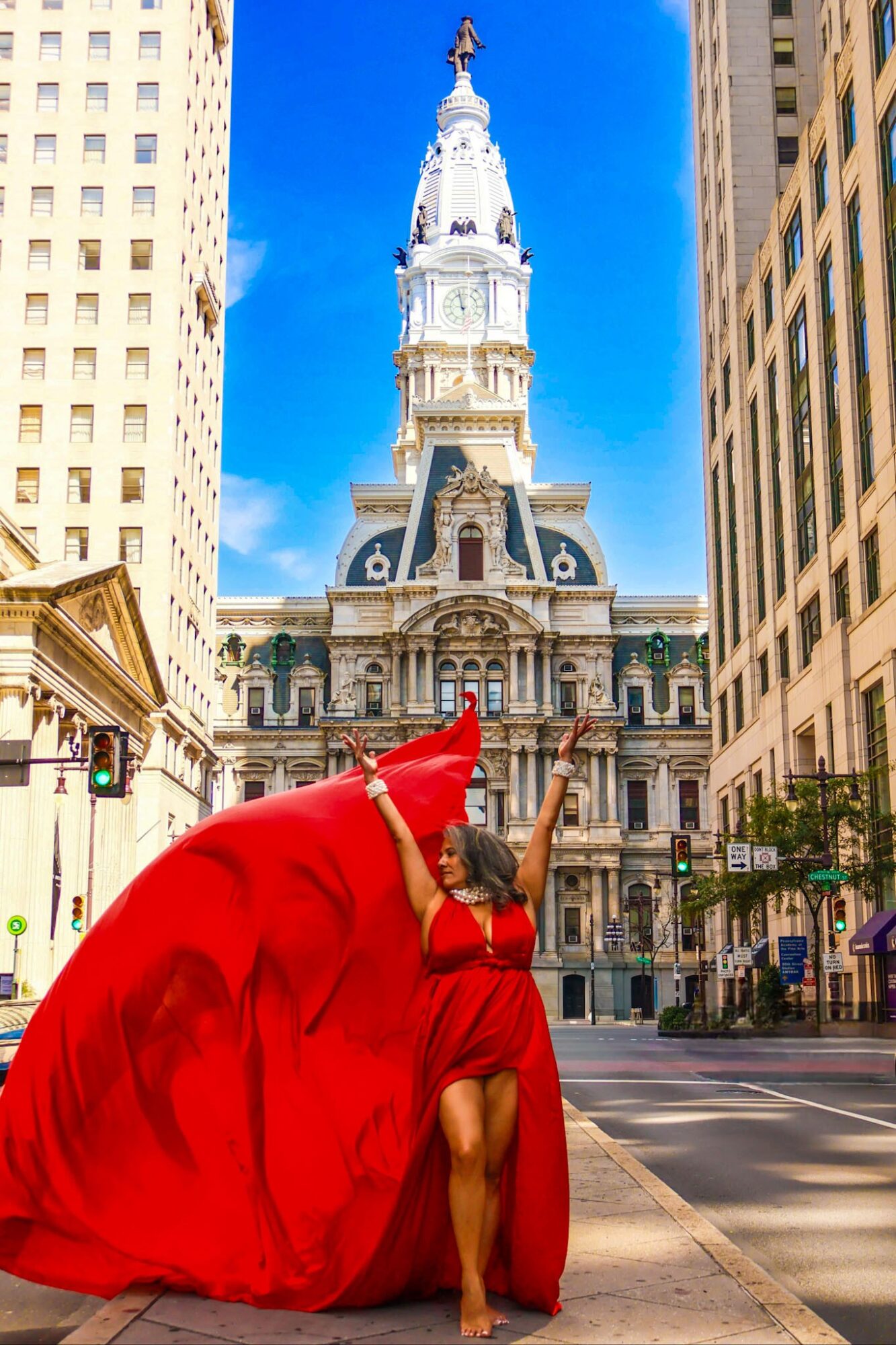 Woman in red dress dancing on city street with historic building and clock tower behind her.