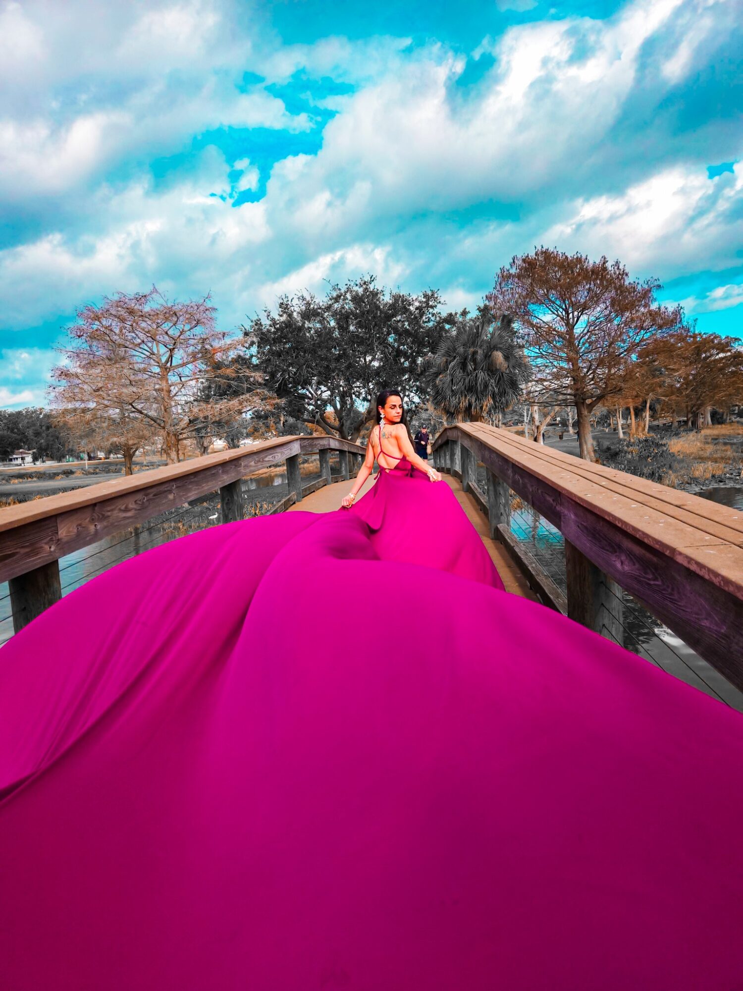 Woman in a bright pink gown standing on a bridge outdoors with trees and a cloudy sky in the background.