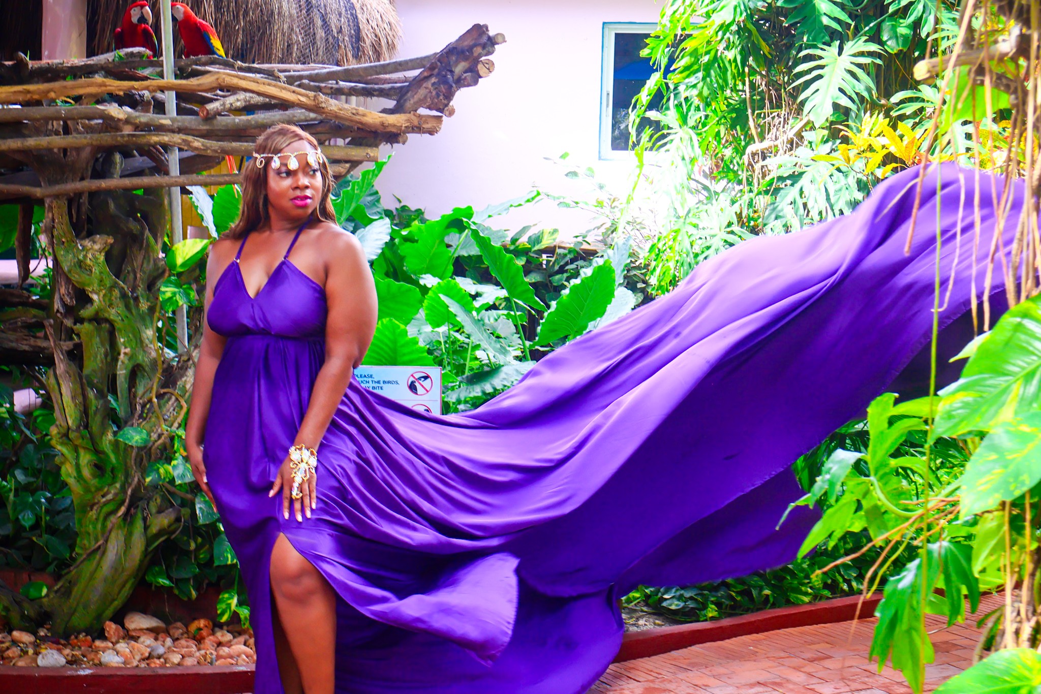 Woman in a purple dress standing outdoors with lush green plants and trees, flowing fabric extending behind her, bright natural light.