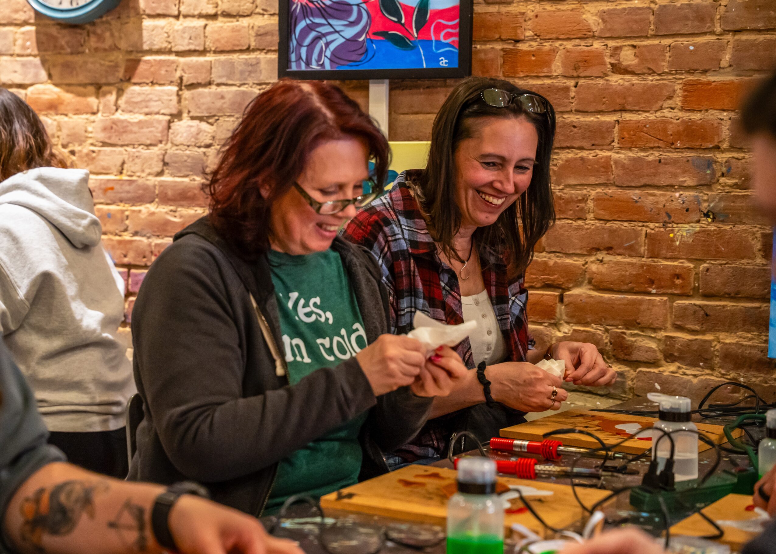 Two women smiling and working with electronic tools at a table in a brick-walled room.