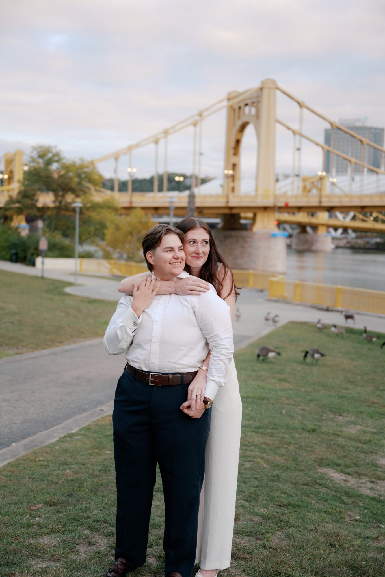 Two women standing on a grassy area near a river with a large bridge in the background, one hugging the other.